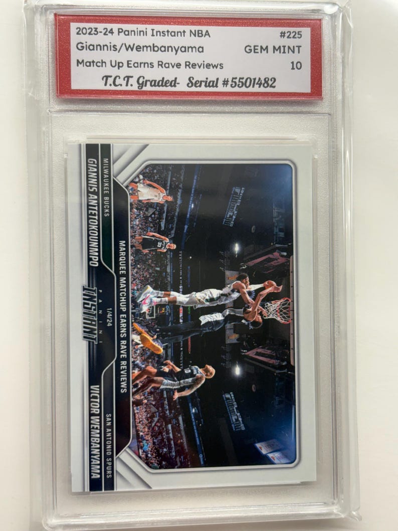 May include: A basketball trading card featuring Giannis Antetokounmpo and Victor Wembanyama. The card is graded GEM MINT 10 by T.C.T. and is part of the 2023-24 Panini Instant NBA series. The card's title is "Match Up Earns Rave Reviews."