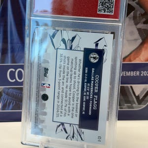 May include: A basketball trading card in a clear protective case. The card details Cooper Flagg, a Dallas Mavericks forward, with his height, weight, and shooting stats. The card is presented on a clear plastic stand.