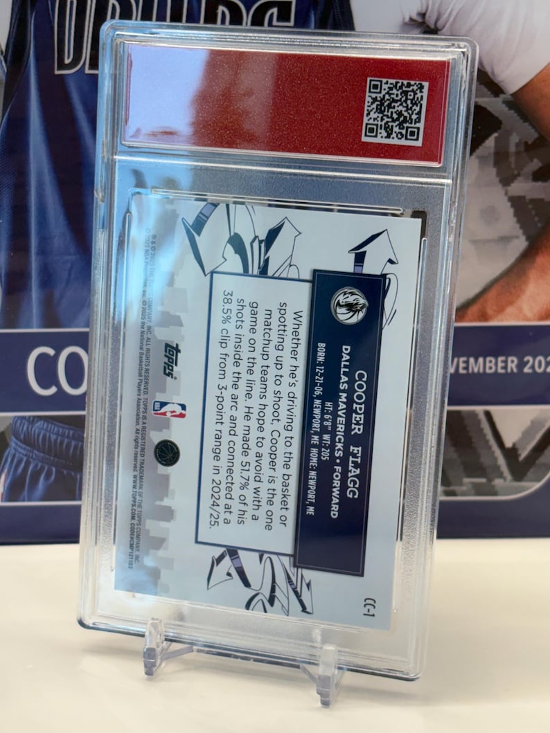 May include: A basketball trading card in a clear protective case. The card details Cooper Flagg, a Dallas Mavericks forward, with his height, weight, and shooting stats. The card is presented on a clear plastic stand.