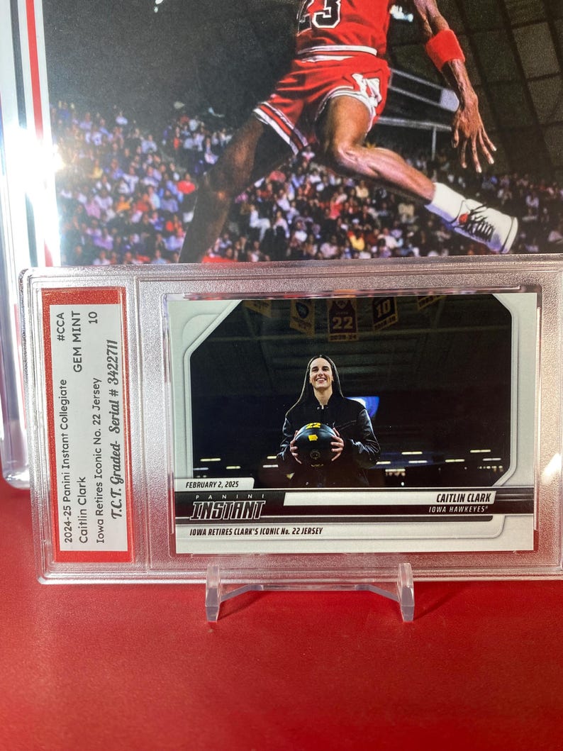 May include: A graded trading card of Caitlin Clark, a basketball player for the Iowa Hawkeyes. The card is from the 2024-25 Panini Instant Collegiate series and features a photo of Clark holding a basketball. The card is graded GEM MINT 10 by T.C.T. and has a serial number of 3422711.