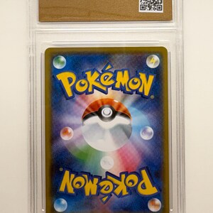 Puede incluir: Una carta coleccionable de Pok&eacute;mon calificada, encerrada en un soporte de pl&aacute;stico transparente. El reverso de la carta presenta un fondo azul y morado con el logotipo de Pok&eacute;mon en amarillo. Un c&oacute;digo QR es visible en la parte superior.