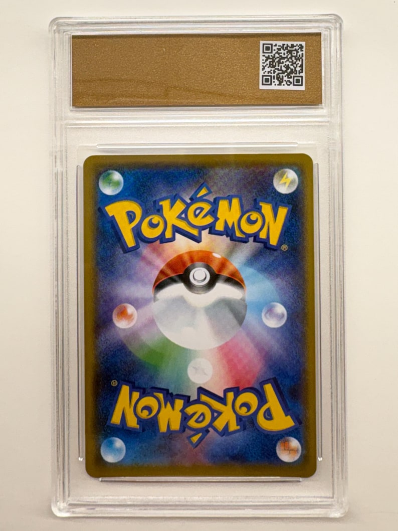 Puede incluir: Una carta coleccionable de Pok&eacute;mon calificada, encerrada en un soporte de pl&aacute;stico transparente. El reverso de la carta presenta un fondo azul y morado con el logotipo de Pok&eacute;mon en amarillo. Un c&oacute;digo QR es visible en la parte superior.