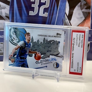May include: A basketball card featuring Cooper Flagg of the Dallas Mavericks, encased for protection. The card displays the text "CLUTCHCity Prospects" and the team logo. It is graded GEM MINT 10.