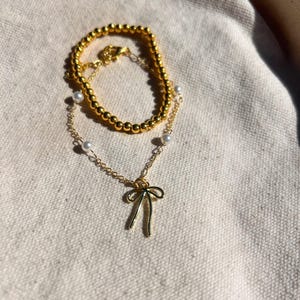 Puede incluir: Un collar de cadena dorada con un delicado colgante de lazo y una pulsera de cuentas doradas. El collar tiene pequeñas perlas blancas espaciadas a lo largo de la cadena.