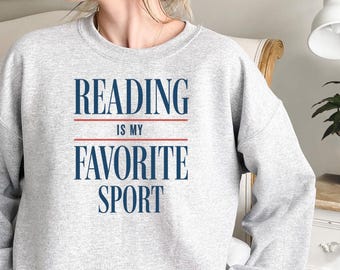 May include: Light gray sweatshirt with the text "READING is my FAVORITE SPORT" in navy blue and red. The sweatshirt is a crew neck style, perfect for casual wear. The image shows a person wearing the sweatshirt.
