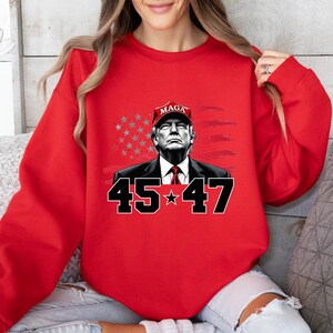 Puede incluir: Sudadera roja con un gr&aacute;fico de un hombre con una gorra "MAGA", con los n&uacute;meros "45" y "47" debajo. El fondo incluye una bandera estadounidense estilizada. La sudadera es de color rojo liso.