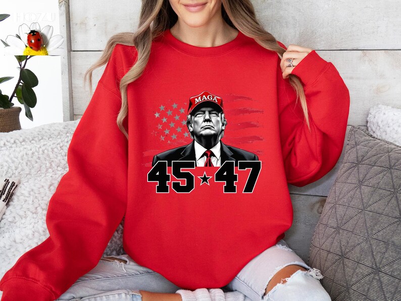 Puede incluir: Sudadera roja con un gr&aacute;fico de un hombre con una gorra "MAGA", con los n&uacute;meros "45" y "47" debajo. El fondo incluye una bandera estadounidense estilizada. La sudadera es de color rojo liso.
