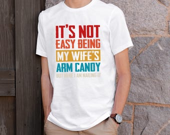 May include: White t-shirt with a vintage-style graphic that reads "It's not easy being my wife's arm candy but here I am nailing it."