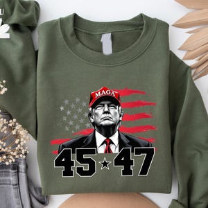 Puede incluir: Sudadera verde oliva con un retrato de un hombre con una gorra roja "MAGA", con la bandera estadounidense de fondo. Los n&uacute;meros "45 * 47" se muestran en blanco y negro.