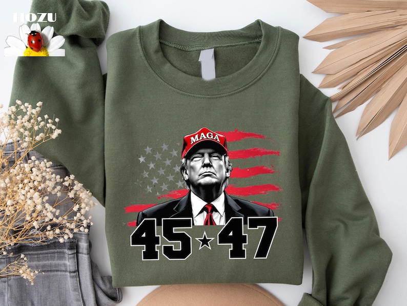 Puede incluir: Sudadera verde oliva con un retrato de un hombre con una gorra roja "MAGA", con la bandera estadounidense de fondo. Los n&uacute;meros "45 * 47" se muestran en blanco y negro.