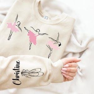 May include: A beige sweatshirt with a black outline of three ballerinas in pink tutus. The word "Christine" is written on the sleeve of the sweatshirt.