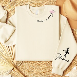 May include: A cream-colored sweatshirt with the text "dance mama" and a silhouette of a ballerina on the left side of the chest. The right sleeve has the text "Hannah" and a silhouette of a ballerina.
