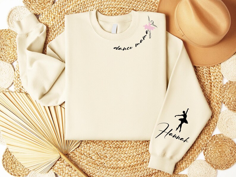 May include: A cream-colored sweatshirt with the text "dance mama" and a silhouette of a ballerina on the left side of the chest. The right sleeve has the text "Hannah" and a silhouette of a ballerina.