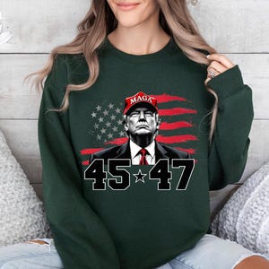 Puede incluir: Sudadera verde oscuro con un gr&aacute;fico de un hombre con una gorra roja "MAGA", sobre un fondo de bandera estadounidense estilizada. Los n&uacute;meros "45" y "47" est&aacute;n debajo, en negrita.