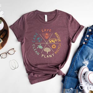 Puede incluir: Una camiseta color marr&oacute;n con un dise&ntilde;o gr&aacute;fico de un c&iacute;rculo con las palabras "Save, Care, Clean, Plant, Rescue, Recycle" alrededor del exterior. Dentro del c&iacute;rculo hay im&aacute;genes de una abeja, una tortuga, un &aacute;rbol, un rinoceronte y un globo terr&aacute;queo.