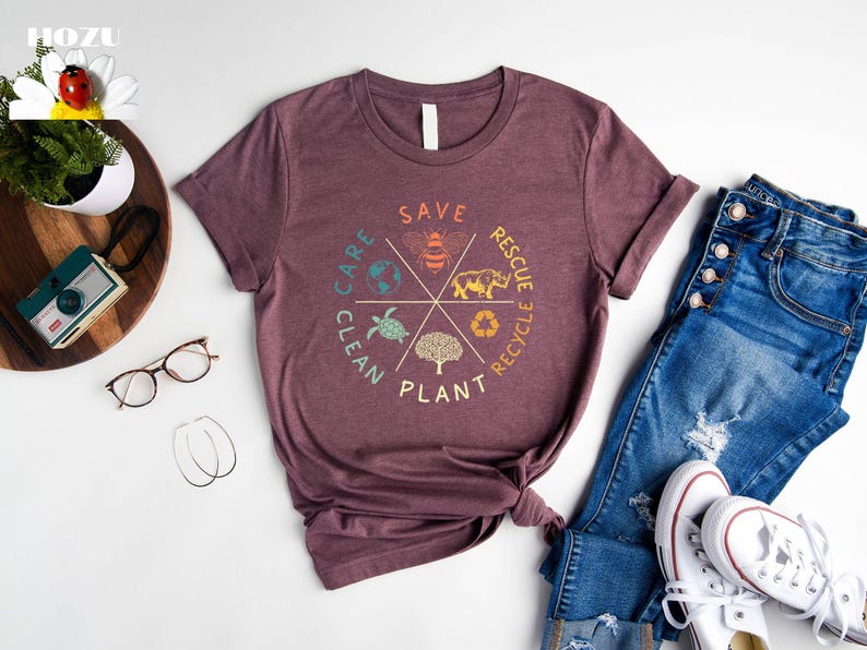 Puede incluir: Una camiseta color marr&oacute;n con un dise&ntilde;o gr&aacute;fico de un c&iacute;rculo con las palabras "Save, Care, Clean, Plant, Rescue, Recycle" alrededor del exterior. Dentro del c&iacute;rculo hay im&aacute;genes de una abeja, una tortuga, un &aacute;rbol, un rinoceronte y un globo terr&aacute;queo.