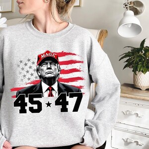 Puede incluir: Sudadera gris claro con un gr&aacute;fico de un hombre con una gorra roja "MAGA", sobre un dise&ntilde;o de bandera estadounidense roja y blanca. El n&uacute;mero "45 * 47" est&aacute; impreso en negro debajo.