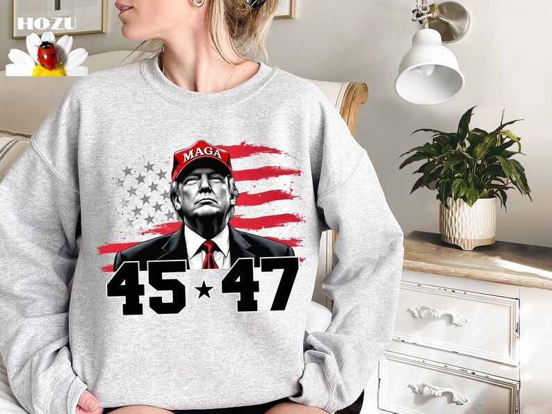 Puede incluir: Sudadera gris claro con un gr&aacute;fico de un hombre con una gorra roja "MAGA", sobre un dise&ntilde;o de bandera estadounidense roja y blanca. El n&uacute;mero "45 * 47" est&aacute; impreso en negro debajo.