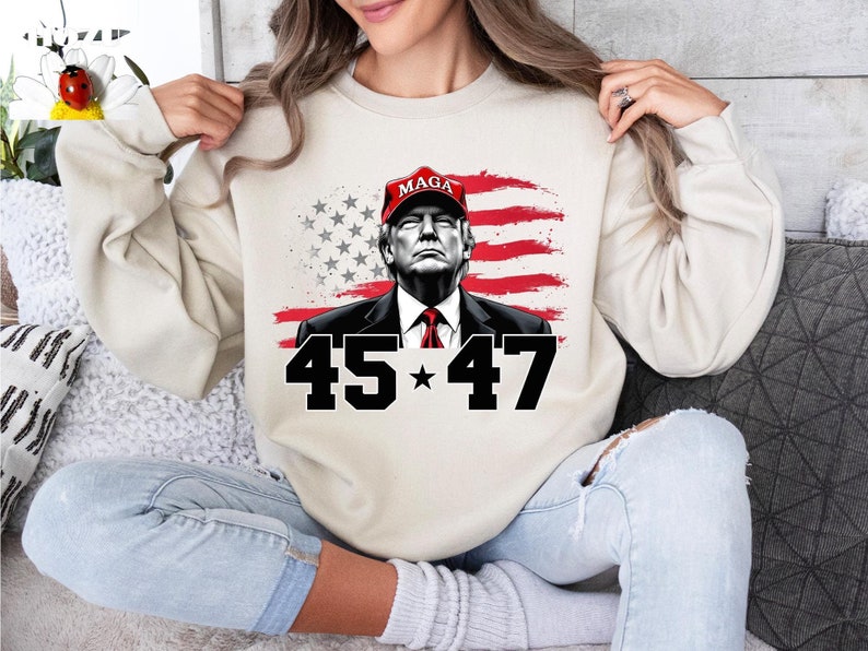 Puede incluir: Sudadera beige con un gr&aacute;fico de un hombre con una gorra roja "MAGA", con la bandera estadounidense de fondo. Los n&uacute;meros "45" y "47" se muestran en negro debajo de la imagen.