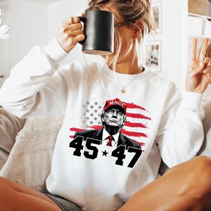 Puede incluir: Sudadera blanca con un gr&aacute;fico de un hombre con una gorra roja "MAGA", con la bandera estadounidense de fondo. Los n&uacute;meros "45" y "47" flanquean una estrella. La sudadera tiene mangas largas. El dise&ntilde;o est&aacute; impreso en la parte delantera.