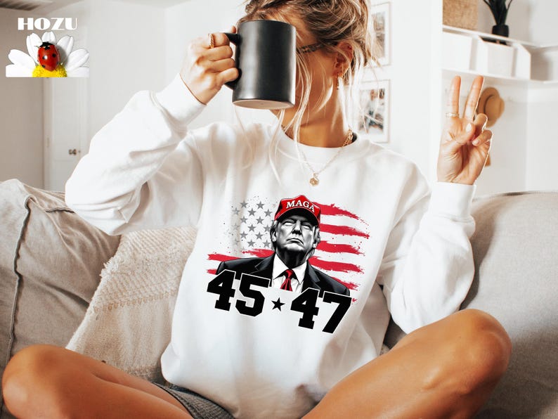 Puede incluir: Sudadera blanca con un gr&aacute;fico de un hombre con una gorra roja "MAGA", con la bandera estadounidense de fondo. Los n&uacute;meros "45" y "47" flanquean una estrella. La sudadera tiene mangas largas. El dise&ntilde;o est&aacute; impreso en la parte delantera.