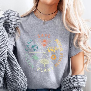Puede incluir: Una camiseta gris con un dise&ntilde;o circular que incluye las palabras "Save, Care, Clean, Plant, Recycle, Reduce" e ilustraciones de una abeja, un &aacute;rbol, un globo terr&aacute;queo, una hoja y un s&iacute;mbolo de reciclaje.