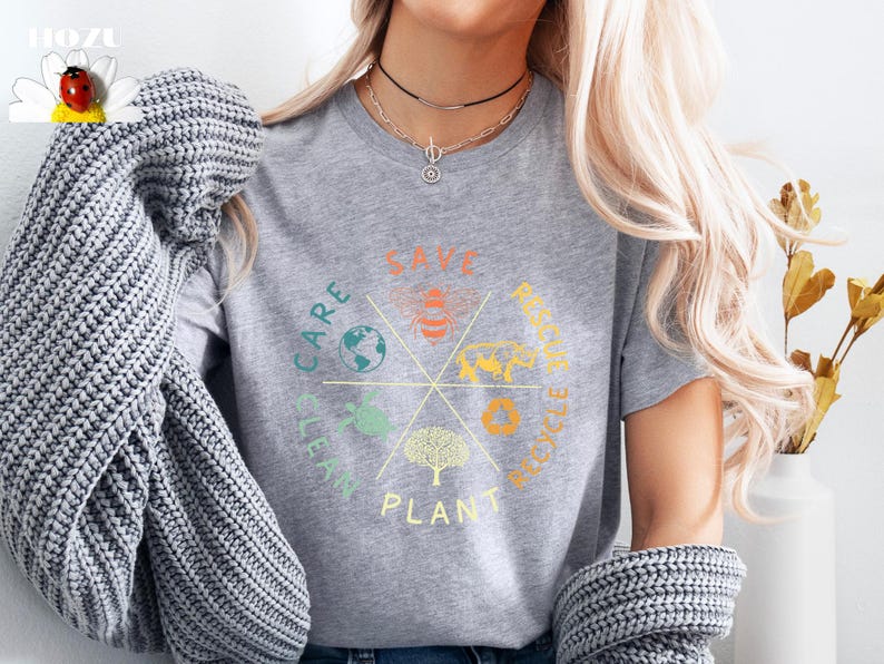 Puede incluir: Una camiseta gris con un dise&ntilde;o circular que incluye las palabras "Save, Care, Clean, Plant, Recycle, Reduce" e ilustraciones de una abeja, un &aacute;rbol, un globo terr&aacute;queo, una hoja y un s&iacute;mbolo de reciclaje.