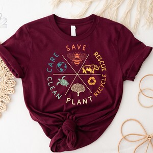 Puede incluir: Una camiseta color burdeos con un dise&ntilde;o gr&aacute;fico de un c&iacute;rculo con las palabras "Save, Plant, Clean, Care, Recycle, Rescue" alrededor del exterior. Dentro del c&iacute;rculo hay im&aacute;genes de una abeja, un rinoceronte, una tortuga, un &aacute;rbol, un globo terr&aacute;queo y un s&iacute;mbolo de reciclaje.