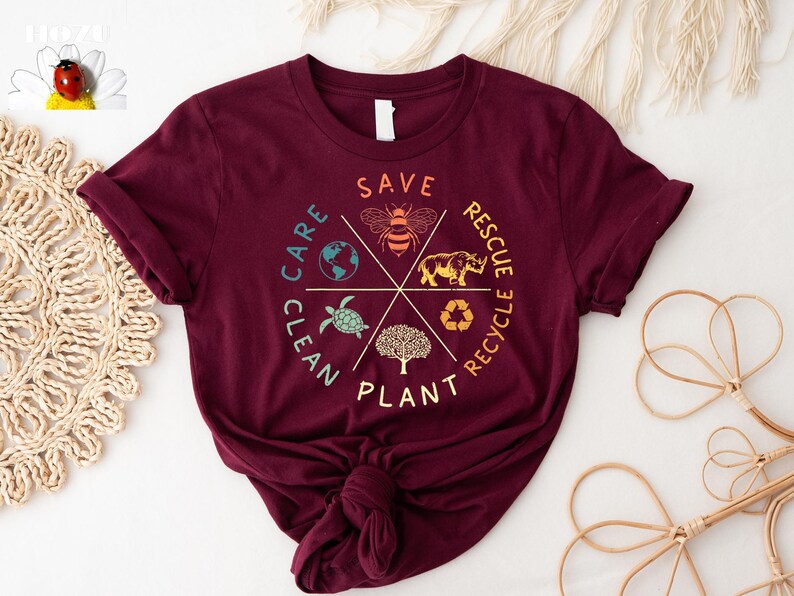 Puede incluir: Una camiseta color burdeos con un dise&ntilde;o gr&aacute;fico de un c&iacute;rculo con las palabras "Save, Plant, Clean, Care, Recycle, Rescue" alrededor del exterior. Dentro del c&iacute;rculo hay im&aacute;genes de una abeja, un rinoceronte, una tortuga, un &aacute;rbol, un globo terr&aacute;queo y un s&iacute;mbolo de reciclaje.
