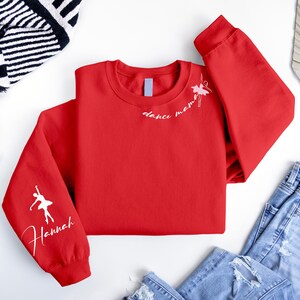 May include: Red sweatshirt with white text that says "dance mommy" and a white silhouette of a ballerina on one sleeve. The other sleeve has the name "Hannah" written in white.