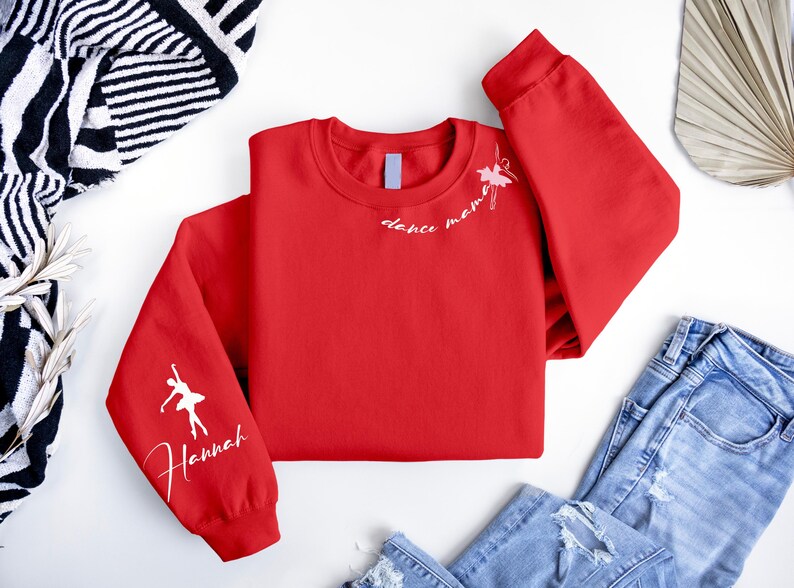 May include: Red sweatshirt with white text that says "dance mommy" and a white silhouette of a ballerina on one sleeve. The other sleeve has the name "Hannah" written in white.
