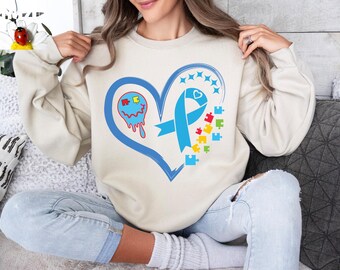 Puede incluir: Una sudadera de color crema con un diseño de corazón azul. El corazón presenta una cinta azul, una pieza de rompecabezas azul y una pieza de rompecabezas azul y roja con una cara. La sudadera está siendo usada por una persona.