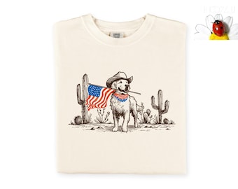 Puede incluir: Camiseta color crema con un dibujo de un golden retriever con sombrero de vaquero y pañuelo, sosteniendo una bandera americana. El perro está situado frente a un paisaje desértico con cactus.