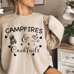 May include: A beige sweatshirt with the text "Campfires & Cocktails" in black lettering. The design includes a campfire, a cocktail glass, and hearts.