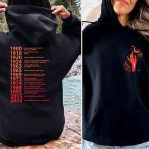 May include: Two black hooded sweatshirts. One features a list of historical events related to women's rights in the United States from 1900 to 2022. The other features a red hand reaching for a crescent moon with stars and leaves.