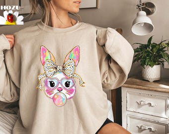 May include: A beige sweatshirt with a colorful cartoon bunny wearing heart-shaped sunglasses and a bow. The bunny is holding a pink and white Easter egg.
