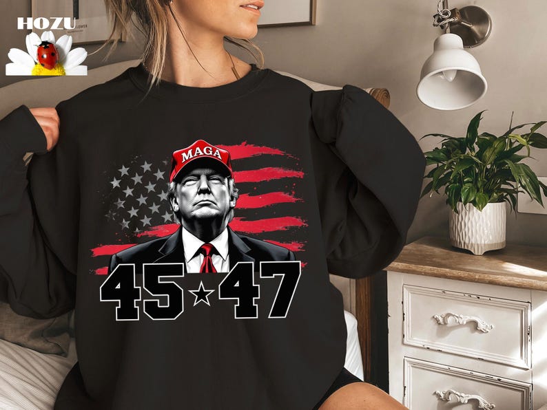Puede incluir: Sudadera negra con un retrato de un hombre con una gorra roja "MAGA", sobre una bandera americana estilizada. El n&uacute;mero "45*47" se muestra debajo en blanco.