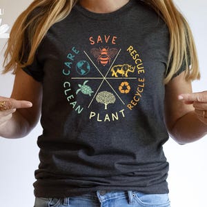 Puede incluir: Una camiseta gris oscuro con un dise&ntilde;o gr&aacute;fico colorido que presenta un c&iacute;rculo con las palabras "Clean, Care, Plant, Save, Rescue, Recycle" e im&aacute;genes de una abeja, una tortuga, un rinoceronte, un &aacute;rbol y un globo terr&aacute;queo.