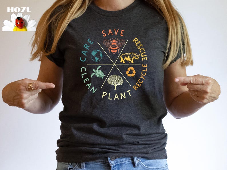 Puede incluir: Una camiseta gris oscuro con un dise&ntilde;o gr&aacute;fico colorido que presenta un c&iacute;rculo con las palabras "Clean, Care, Plant, Save, Rescue, Recycle" e im&aacute;genes de una abeja, una tortuga, un rinoceronte, un &aacute;rbol y un globo terr&aacute;queo.
