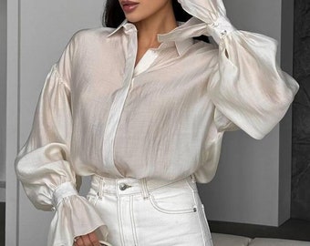 May include: A cream-coloured blouse with long, billowy sleeves and a button-down front is paired with white jeans. A small, light-coloured handbag with silver hardware is held in the hand. The outfit is a monochromatic, elegant style.