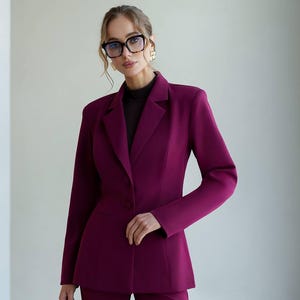 May include: A plum-colored pantsuit featuring a tailored blazer with a two-button closure and matching trousers. The blazer has structured shoulders and a fitted silhouette. The outfit is worn with a dark turtleneck. The person is wearing glasses and gold earrings.
