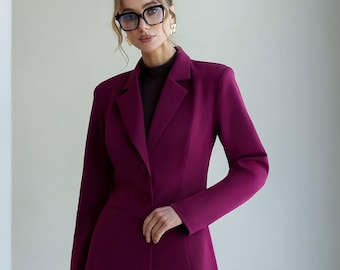 May include: A plum-colored pantsuit featuring a tailored blazer with a two-button closure and matching trousers. The blazer has structured shoulders and a fitted silhouette. The outfit is worn with a dark turtleneck. The person is wearing glasses and gold earrings.