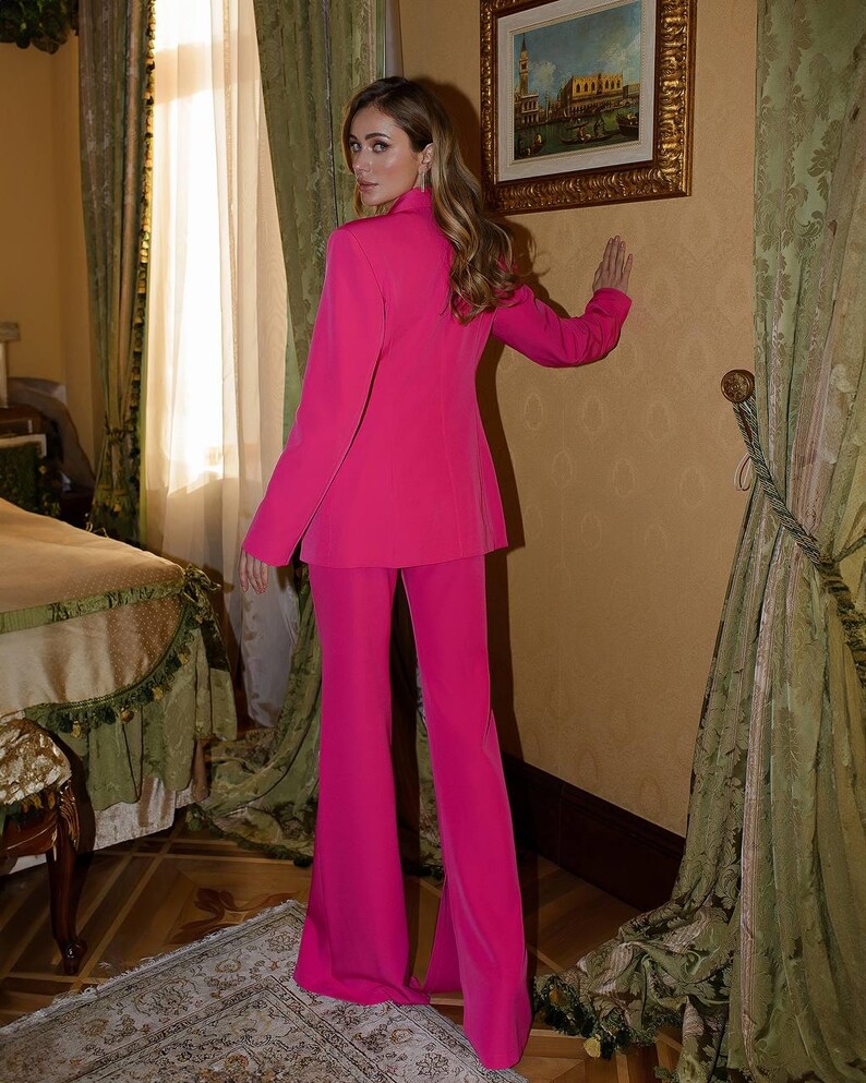 May include: A woman wearing a bright pink pantsuit. The suit features a blazer with a notched collar and a pair of wide-leg pants.
