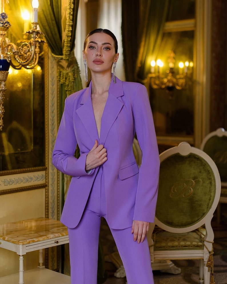 May include: A woman wearing a lavender-colored pantsuit. The suit has a blazer with a notched lapel and a single button closure. The pants are tailored and have a straight leg.