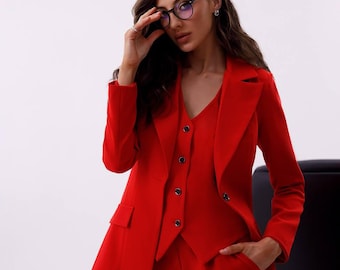 May include: A woman wearing a red suit with a blazer, vest, and pants. She is wearing round glasses and has long brown hair.