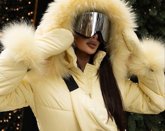Puede incluir: Traje de esquí amarillo claro con capucha forrada de piel y guantes a juego. El traje presenta una hebilla de cinturón plateada y una riñonera a juego. La persona lleva gafas de esquí plateadas.