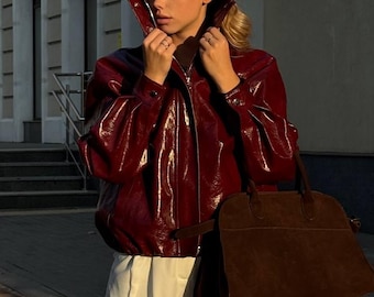 May include: A woman wearing a shiny, burgundy-colored jacket with a high collar and a brown suede handbag. She is also wearing white shorts. The jacket has a zipper closure and a glossy finish. The handbag has a structured shape.