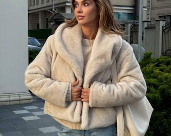 May include: A beige faux fur jacket with a wide collar and a matching sweater. The jacket is paired with light blue jeans and a cream-colored tote bag. The outfit is worn outdoors on a city street.