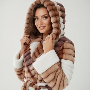 May include: A multi-toned hooded coat in shades of brown, white, and tan. The coat has a textured, layered pattern and white cuffs. The model is wearing the coat and smiling.