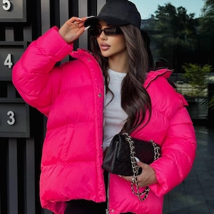 Peut inclure: Une doudoune rose vif, une casquette de baseball noire et des lunettes de soleil. La veste a une fermeture éclair et un motif matelassé. Un sac à main noir matelassé avec une bandoulière en chaîne dorée est également visible.